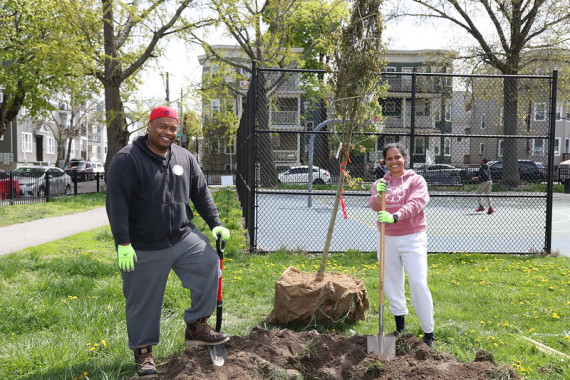 Two volunteers standing around a hole with a tree in the background that they're about to plant.
